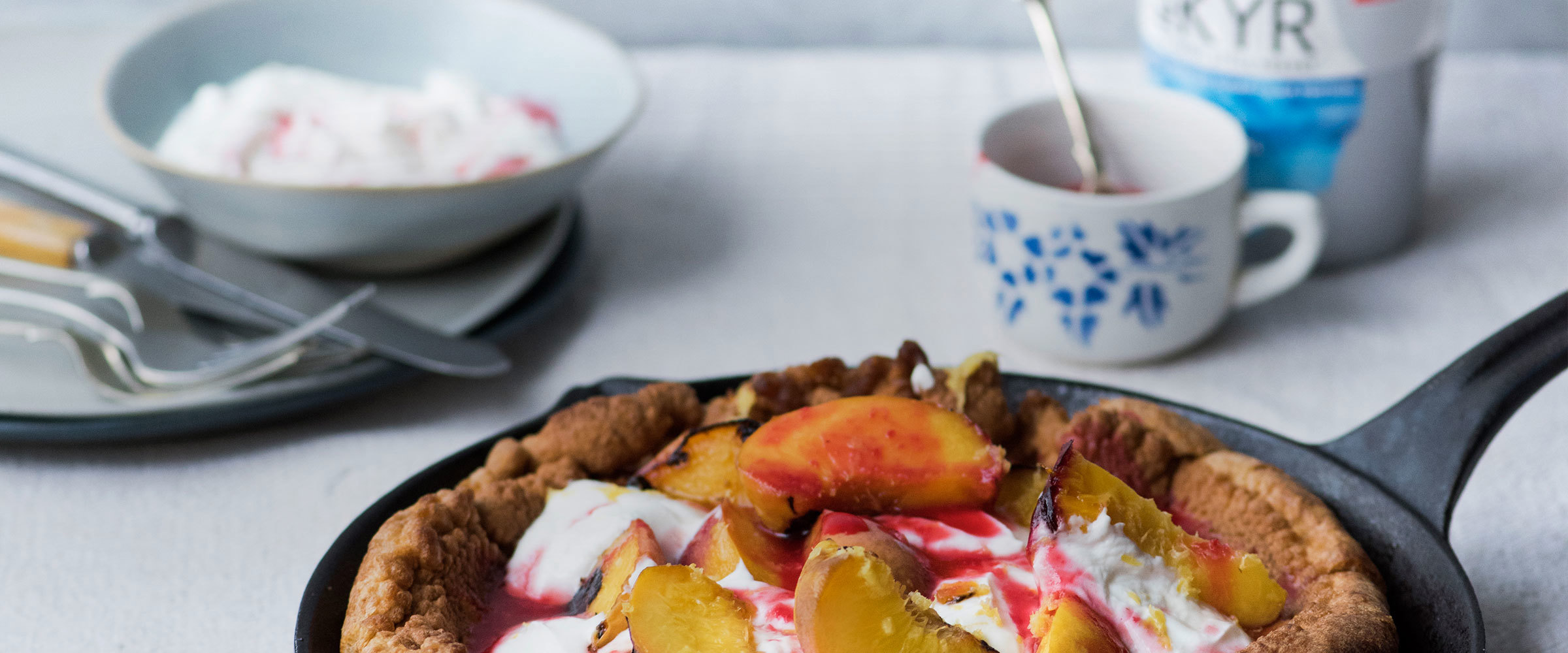 A skillet with a Dutch Baby peach pastry desert on a table with a pot Arla Skyr in the background